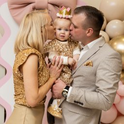 Karina Zioła Fotografia - Uśmiechnięta roczna dziewczynka w koronie, trzymana przez rodziców w złotych strojach, całowana z obu stron na tle dekoracji z balonów i kokardy.