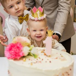 Karina Zioła Fotografia - Uśmiechnięta roczna dziewczynka w koronie z cyfrą '1' przy torcie urodzinowym, obok brat w muszce i dłoń dorosłego. Jasne, ciepłe światło, pastelowe dekoracje.