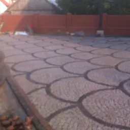 Ułożona nawierzchnia z kostki granitowej w kształcie łusek, widoczne prace wykończeniowe i fragment muru w tle.