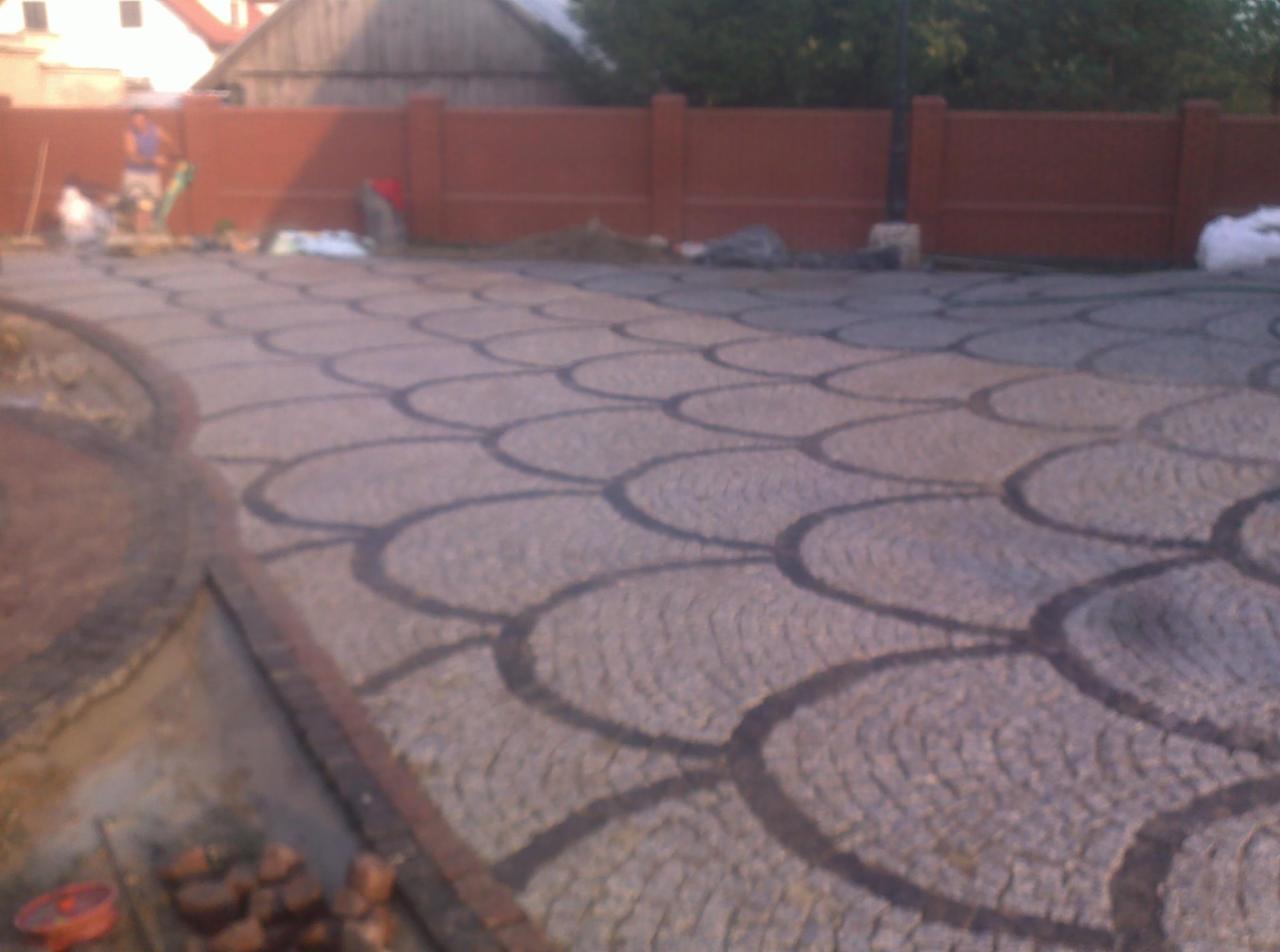 Ułożona nawierzchnia z kostki granitowej w kształcie łusek, widoczne prace wykończeniowe i fragment muru w tle.