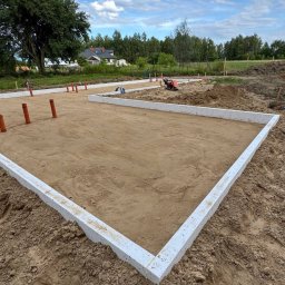 VLADIS Vladyslav Rybachuk - Prostokątny obrys betonowych ław fundamentowych na wyrównanym piaszczystym podłożu, z wystającymi pomarańczowymi rurami instalacyjnymi. W tle dom i zieleń.