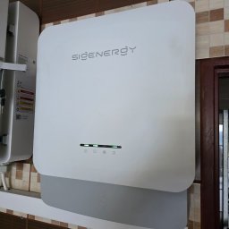 LILIANNA SOŁTAN Step by lili - Biały magazyn energii SIGENERGY zamontowany na ścianie z brązowymi i beżowymi płytkami. Widoczne diody LED i etykiety ostrzegawcze. Częściowo widoczna rama drzwi.