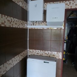 LILIANNA SOŁTAN Step by lili - Biały magazyn energii Side Energy zamontowany na brązowo-beżowej ścianie z płytek, obok drzwi. Widoczne przewody i skrzynka elektryczna. Wnętrze pomieszczenia.