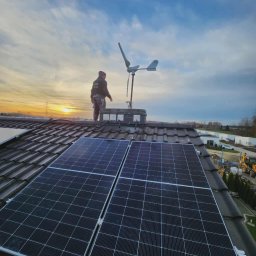 LILIANNA SOŁTAN Step by lili - Instalacja paneli słonecznych i małej turbiny wiatrowej na dachu domu jednorodzinnego o zmierzchu. Widoczny monter i elementy instalacji.