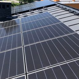 LILIANNA SOŁTAN Step by lili - Panele fotowoltaiczne na dachu z czarną dachówką, widok z góry, komin z cegły w tle, instalacja solarna na ukończonym dachu, nowoczesne rozwiązanie.
