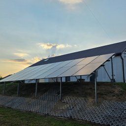 LILIANNA SOŁTAN Step by lili - Naziemna instalacja paneli słonecznych na pochyłym, trawiastym terenie z betonowymi kratami, obok nowoczesnego domu z ciemnym dachem, w ciepłym świetle zachodzącego słońca.