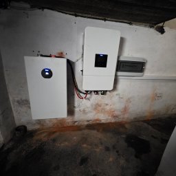 LILIANNA SOŁTAN Step by lili - Nowoczesny biały magazyn energii z okrągłym wyświetlaczem i inwerter z prostokątnym, zamontowane na surowej, poplamionej ścianie piwnicy obok rozdzielnicy.