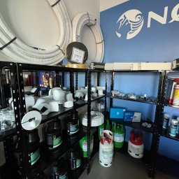 INSTALBUD SYSTEM - Regał z akcesoriami instalacyjnymi: rury, kształtki, środki czyszczące, butle z gazem. Widoczne logo firmy na ścianie. Elementy do instalacji klimatyzacji i wentylacji.
