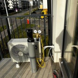 INSTALBUD SYSTEM - Biały klimatyzator zewnętrzny na balkonie, obok butla z gazem i narzędzia. Widoczne przewody w ścianie. W tle zabudowa miejska i żółta ciężarówka.