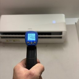 INSTALBUD SYSTEM - Pomiar temperatury klimatyzatora ściennego za pomocą termometru laserowego. Wyświetlacz termometru wskazuje 49 stopni Celsjusza. Biała obudowa klimatyzatora.