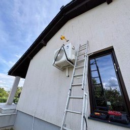 INSTALBUD SYSTEM - Biała jednostka zewnętrzna klimatyzacji zawieszona na ścianie domu, zabezpieczona pasami. Obok drabina oparta o ścianę, narzędzia na parapecie okna.