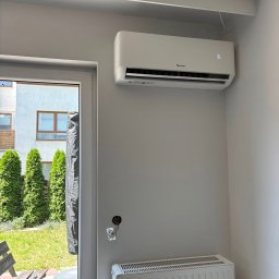 INSTALBUD SYSTEM - Biały klimatyzator NORDIS na szarej ścianie w jasnym wnętrzu, widok z boku. Obok okno z widokiem na zieleń i biały budynek. Grzejnik pod klimatyzatorem.