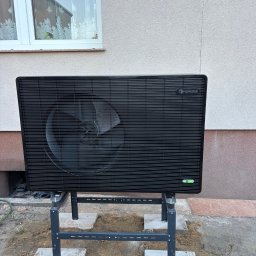 INSTALBUD SYSTEM - Czarna pompa ciepła NORDIS na metalowym stelażu, ustawiona na betonowych płytach przed jasnym budynkiem z oknem. Widoczne elementy instalacji i otoczenie.