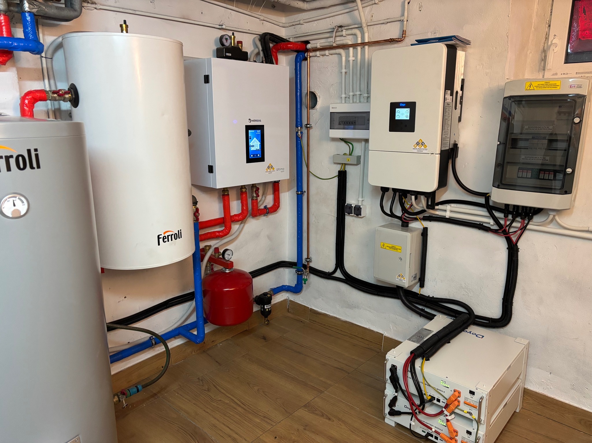 Kompletna instalacja grzewcza i fotowoltaiczna w kotłowni: zbiornik Ferroli, inwerter Nordis Ultima, akumulatory Deye, rozdzielnica elektryczna. Rury w kolorach czerwonym i niebieskim.