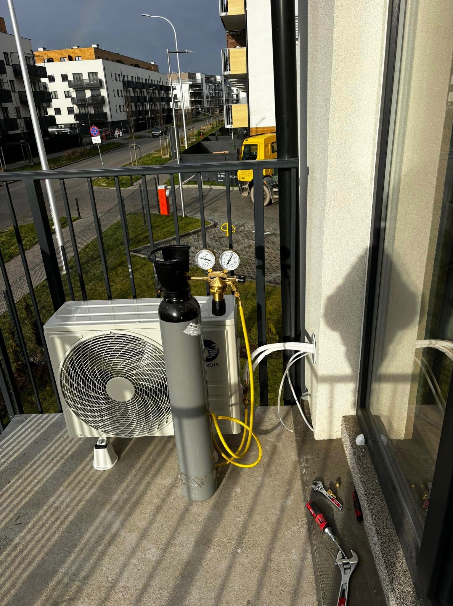 Biały klimatyzator zewnętrzny na balkonie, obok butla z gazem i narzędzia. Widoczne przewody w ścianie. W tle zabudowa miejska i żółta ciężarówka.