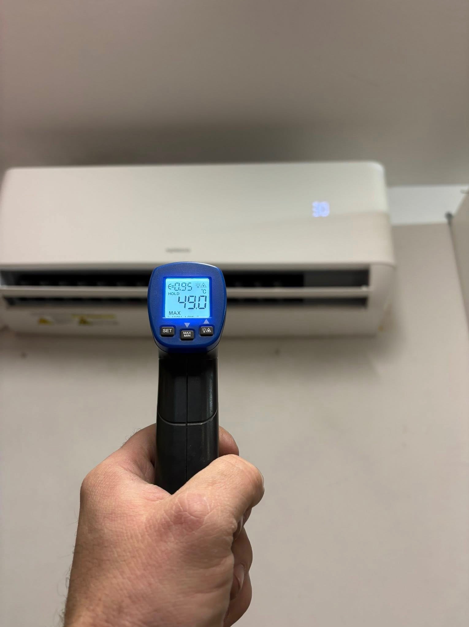 Pomiar temperatury klimatyzatora ściennego za pomocą termometru laserowego. Wyświetlacz termometru wskazuje 49 stopni Celsjusza. Biała obudowa klimatyzatora.