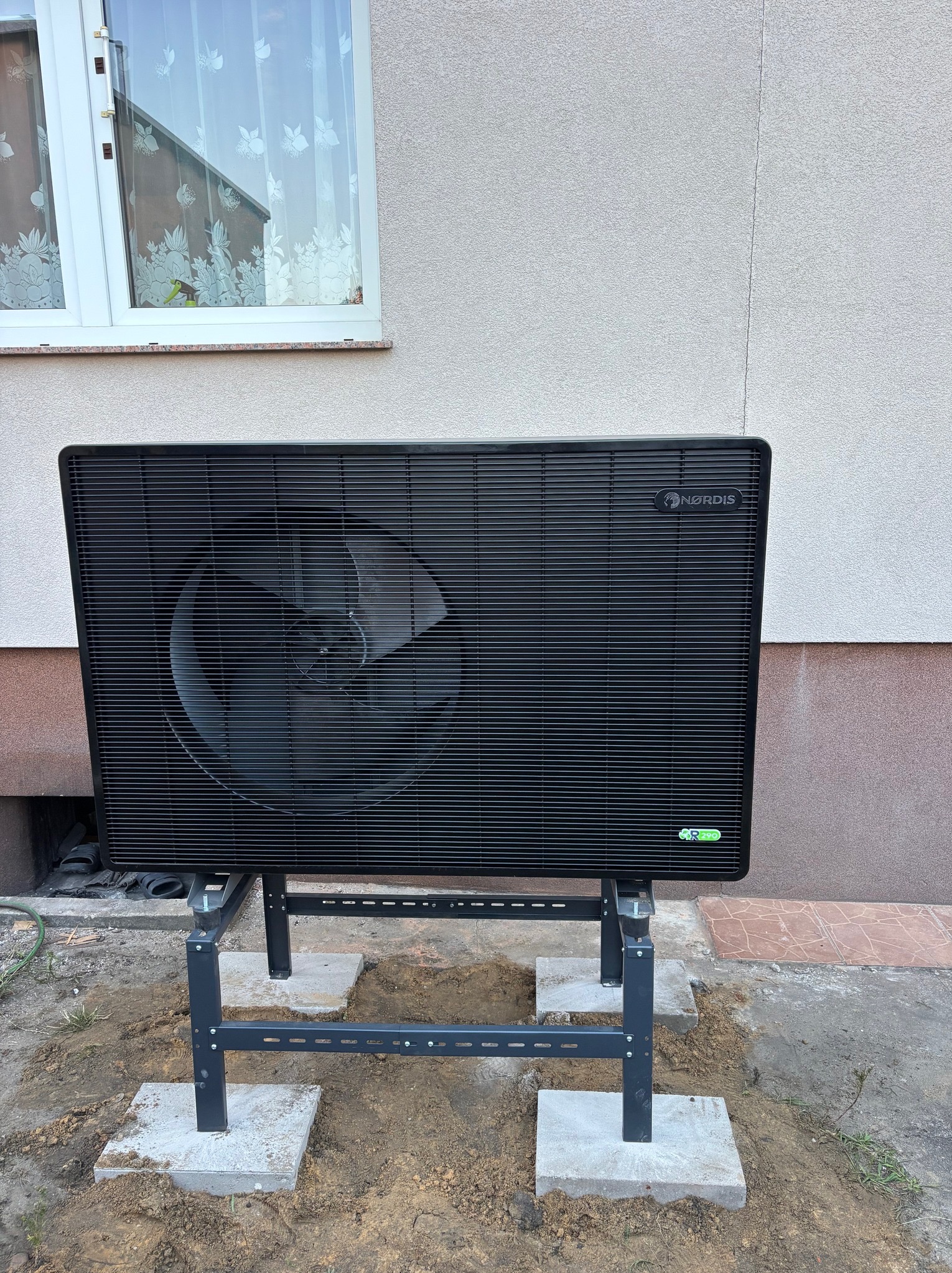 Czarna pompa ciepła NORDIS na metalowym stelażu, ustawiona na betonowych płytach przed jasnym budynkiem z oknem. Widoczne elementy instalacji i otoczenie.