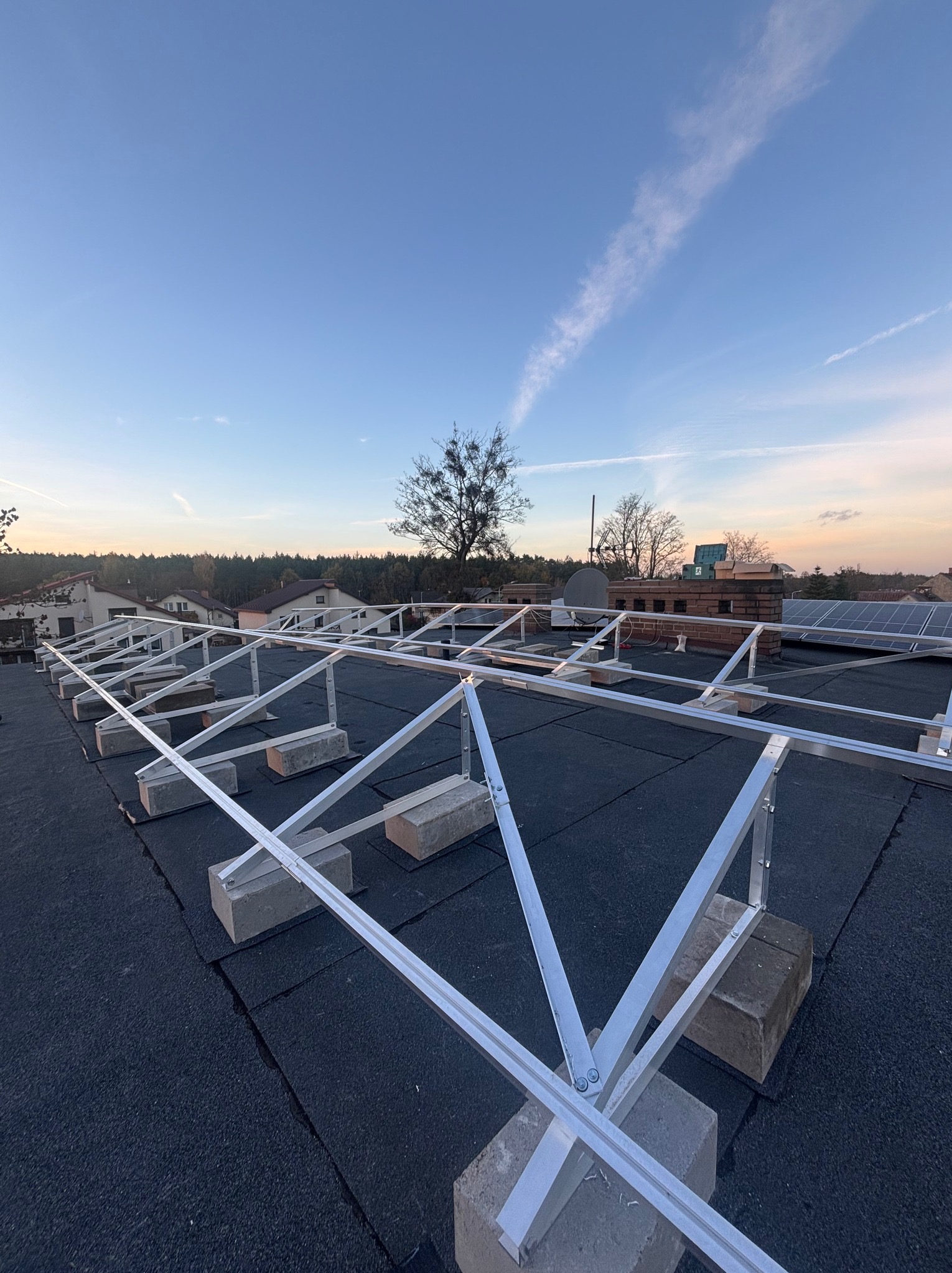Montaż stelażu pod panele fotowoltaiczne na dachu pokrytym papą, widok z góry na konstrukcję z aluminium, w tle zabudowa jednorodzinna i błękitne niebo o zmierzchu.