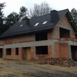 Surowy stan dwukondygnacyjnego domu z cegły, z dachem pokrytym czarną dachówką i widocznymi oknami dachowymi, otoczonego drzewami i z kupką cegieł przed budynkiem.