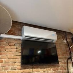 Klima Go - Biały klimatyzator ścienny zamontowany nad telewizorem na tle ceglanej ściany z widocznym fragmentem okrągłej lampy na suficie.