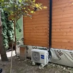 Klima Go - Biała jednostka zewnętrzna klimatyzacji Haier zamontowana przy drewnianym ogrodzeniu, widoczna z boku, z drzewem w tle. Montaż w ogrodzie.