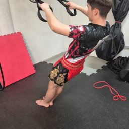 BartDrwal - Mężczyzna w sportowych szortach i koszulce wykonuje ćwiczenie na zawieszeniu TRX w siłowni. Widoczne maty, lina do ćwiczeń i worki treningowe.