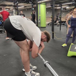BartDrwal - Mężczyzna w białej koszulce opiera się na sztandze w siłowni, w tle inni ćwiczący i sprzęt do ćwiczeń. Widoczne buty sportowe Adidas.