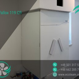Eco team Łukasz Kulaszewski - Biała jednostka rekuperacyjna Vallox 119 Cfi zamontowana w pomieszczeniu, widoczne elementy instalacji i okno w tle. Ujęcie z dołu, z logo firmy w rogu.