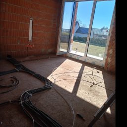 MA-Elektro - Wnętrze w stanie surowym z widoczną instalacją elektryczną w peszlach na podłodze, ściany z cegły dziurawki, okno z widokiem na domy.