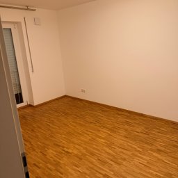 Górale Transport sp. z o.o. - Pusty pokój z drewnianą podłogą i białymi ścianami, oświetlony lampą sufitową. Widok zza uchylonych drzwi, ukazujący przygotowanie do przeprowadzki lub opróżniania.