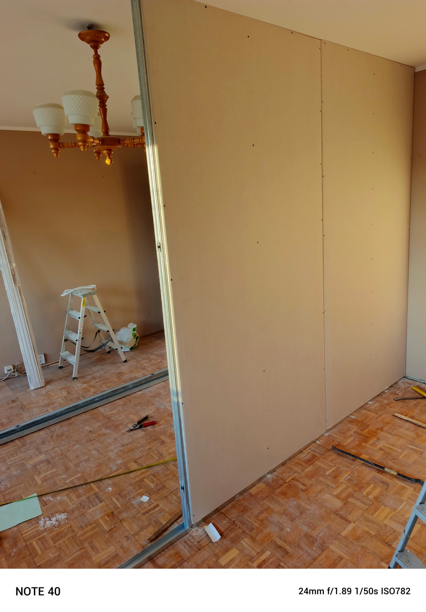 Montaż płyt kartonowo-gipsowych na metalowym stelażu w trakcie remontu pokoju, widoczna drabina, narzędzia i resztki materiałów na podłodze z parkietu.