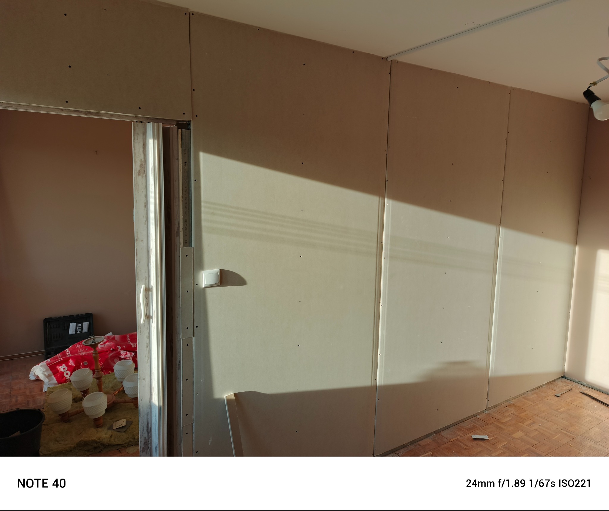 Wnętrze w trakcie remontu: ściany wyłożone płytami gipsowo-kartonowymi, widoczne ślady po wkrętach, fragment podłogi z parkietu, w tle przez otwarte drzwi widoczny fragment pomieszczenia...