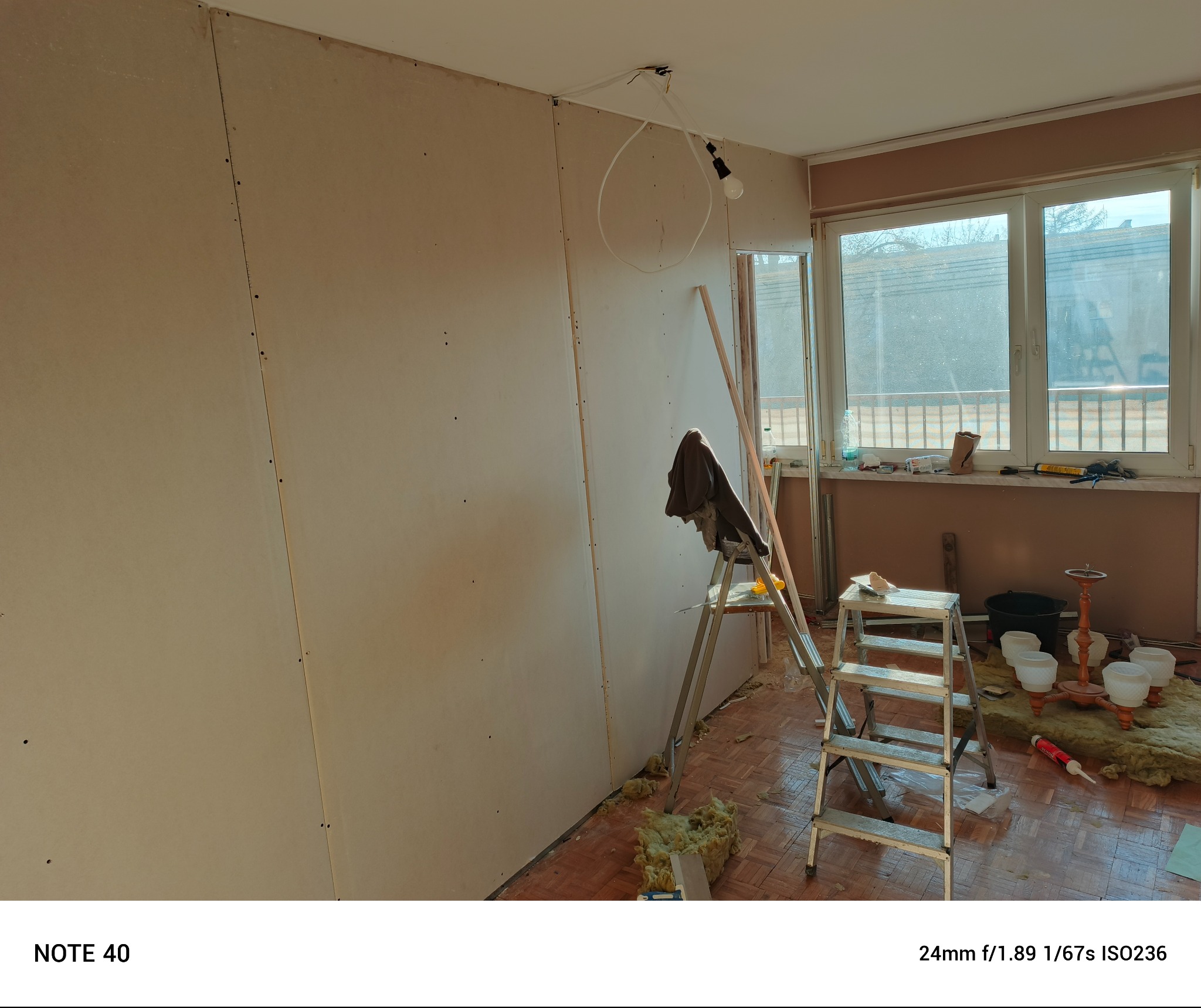 Montaż płyt gipsowo-kartonowych na ścianie, widoczna drabina, lampa wisząca z żarówką i materiały izolacyjne na podłodze w remontowanym pokoju z oknem.