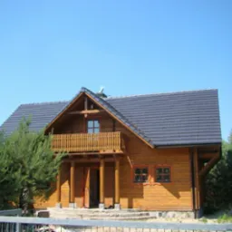 Drewniany dom szkieletowy z balkonem i ciemną dachówką, otoczony zielenią, widziany zza metalowego ogrodzenia w słoneczny dzień. Charakterystyczna konstrukcja.