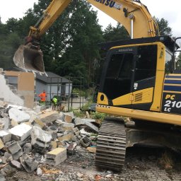 KONEKT - Żółta koparka Komatsu PC210 LC z łyżką nad stertą gruzu po rozbiórce budynku. W tle pracownicy w odblaskowych ubraniach i częściowo zburzona ściana.