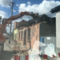 KONEKT - Pomarańczowy dźwig Atlas burzy ceglany budynek z czarnym dachem na tle błękitnego nieba z chmurami. Gruz leży na ulicy. Widoczny znak drogowy i fragment innego budynku.