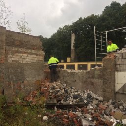 KONEKT - Dwóch pracowników w jaskrawych kamizelkach na tle częściowo zburzonej, ceglano-betonowej ściany. Gruz i drabina w kadrze. W tle budynek i drzewa.