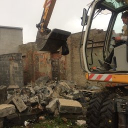 KONEKT - Żółta koparka z operatorem w trakcie rozbiórki ceglanego budynku. Gruz i fragmenty ścian leżą na ziemi. Widoczne detale kabiny operatora i ramię koparki.