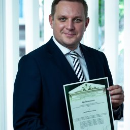 Kancelaria Adwokacka Adw. Karol Niewiarowski - Adwokat Lublin - Uśmiechnięty adwokat w garniturze prezentuje akt ślubowania na tle okna z żaluzjami. Dokument z pieczęcią Okręgowej Rady Adwokackiej w Lublinie.