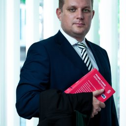 Kancelaria Adwokacka Adw. Karol Niewiarowski - Adwokat Lublin - Portret mężczyzny w garniturze, trzymającego kodeks 'Prawo Karne' i togę adwokacką na tle okna z żaluzjami. Ujęcie od pasa w górę, neutralny wyraz twarzy.