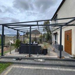 Nowak Okna - Nowoczesna, grafitowa pergola tarasowa z meblami ogrodowymi i roślinami w donicach, przylegająca do domu z brązowymi drzwiami wejściowymi. Uwagę przykuwa piesek.