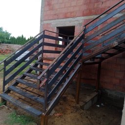 Luka-spaw - Zewnętrzne, dwubiegowe schody metalowe z prostymi balustradami, zamontowane przy budynku z czerwonych pustaków ceramicznych. Widoczne surowe wykończenie i otoczenie placu budowy.