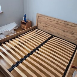 Miłosz - Rama łóżka z drewnianym wezgłowiem i listwami, częściowo zmontowana w pokoju z drewnianą podłogą. Obok szafka nocna i kartony.