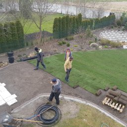 Niwińska Sarkiewicz Ogrody i Wnętrza - Montaż trawy z rolki w ogrodzie z widokiem z góry. Pracownicy rozkładają trawę, widoczne narzędzia i paleta z rolkami trawy. Częściowo ułożona kostka brukowa.