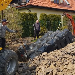 Niwińska Sarkiewicz Ogrody i Wnętrza - Montaż zbiornika retencyjnego w ziemi z użyciem koparki i miniładowarki. Trzech mężczyzn nadzoruje proces. W tle dom z czerwoną dachówką i zielone tuje.