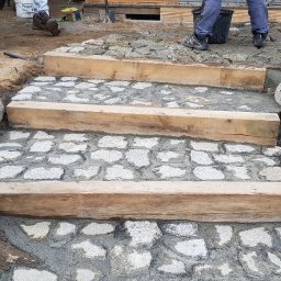 Niwińska Sarkiewicz Ogrody i Wnętrza - Schody z kostki granitowej z drewnianymi belkami, wkomponowane w naturalny krajobraz z drzewami i kamieniami. Etap prac w ogrodzie.