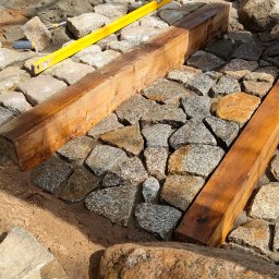 Niwińska Sarkiewicz Ogrody i Wnętrza - Układanie kostki granitowej w ogrodzie, widoczne narzędzia, poziomica i drewniane belki. Częściowo ułożona nawierzchnia z kostki. Prace w toku, etap pośredni.