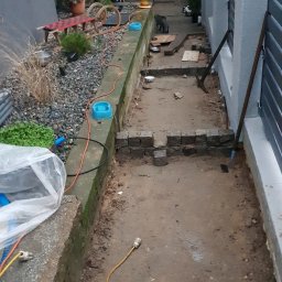 Niwińska Sarkiewicz Ogrody i Wnętrza - Etap układania kostki granitowej w wąskim przejściu ogrodu. Widoczne narzędzia, częściowo ułożona kostka i kot obserwujący prace.