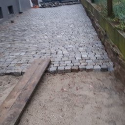 Niwińska Sarkiewicz Ogrody i Wnętrza - Fragment ułożonej kostki granitowej w odcieniach szarości, widoczna deska i podłoże przed dalszym etapem prac. W tle parasol i opona.