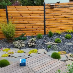 Niwińska Sarkiewicz Ogrody i Wnętrza - Ogród z drewnianym płotem, kamieniami, roślinami i systemem nawadniania Hunter. Widoczny mężczyzna w stroju roboczym. Ujęcie z poziomu podłogi tarasu.